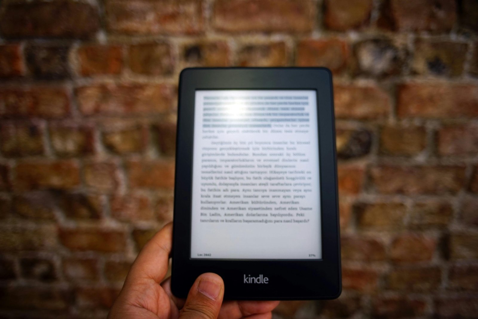 Home Close-up of a Kindle e-reader held by a person, showcasing a digital reading device against a textured brick wall backdrop.